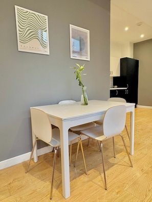 A dining area with a white table, four chairs, a vase with a plant, and two framed artworks on the wall. - Shoreditch Apartments by PrimeCityHosts F4 (Greater London)