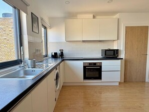 A modern kitchen with a dishwasher, microwave, and a window with a view of a brick building. - Shoreditch Apartments by PrimeCityHosts F7 (Greater London)