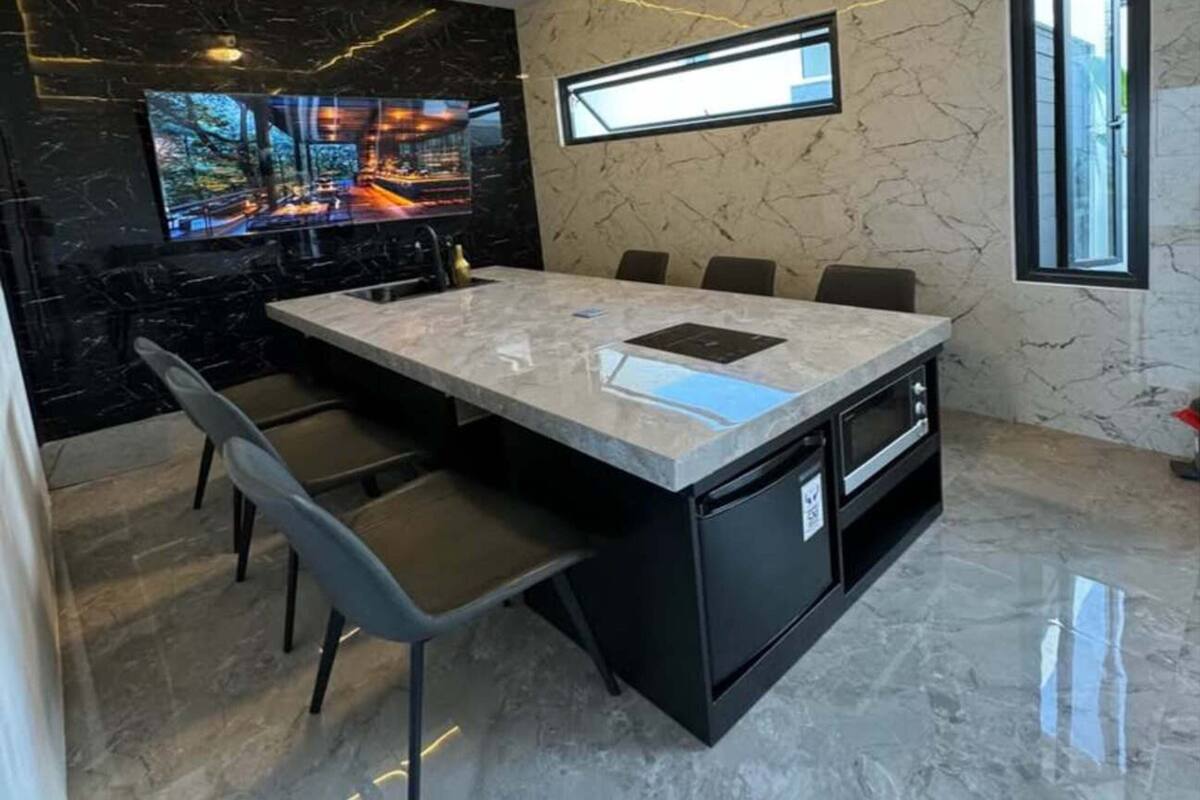 Uma cozinha moderna com uma ilha grande, banquetas pretas, ventilador de teto e uma TV fixada na parede.