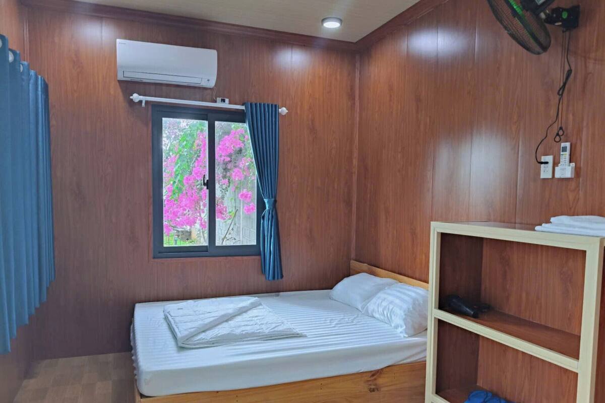 Una habitación pequeña y ordenada con una cama de madera, una ventana con cortinas, un equipo de aire acondicionado y un estante con zapatos.