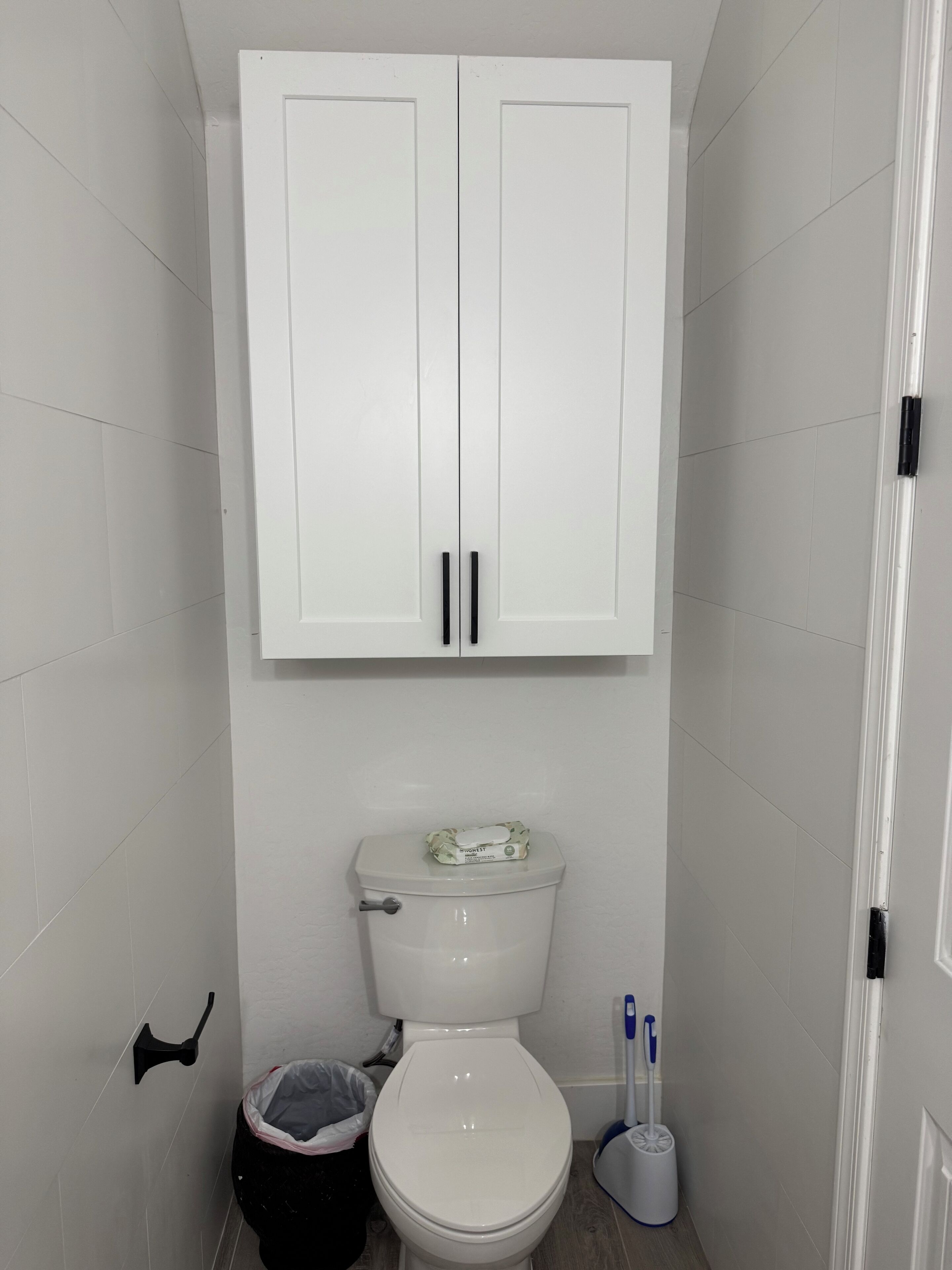 Ein Badezimmer mit Toilette, Mülleimer, Toilettenbürste und einem Schrank über der Toilette.