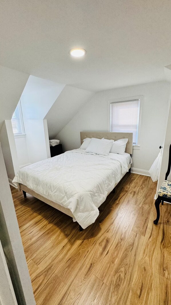 A bedroom with a bed, wooden flooring, a window with blinds, and a sloped ceiling.