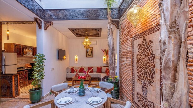 A cozy dining area with a round table set for four, surrounded by wooden chairs. A sofa with red cushions is visible in the background, and a decorative wall with intricate patterns is to the right. - Elegant Riad Musée: Authentic Marrakech Charm in the Medina (marrakech)