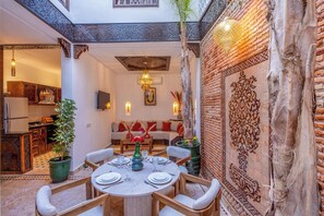 A cozy dining area with a round table set for four, surrounded by wooden chairs. A sofa with red cushions is visible in the background, and a decorative wall with intricate patterns is to the right. - Elegant Riad Musée: Authentic Marrakech Charm in the Medina (marrakech)