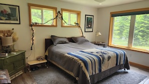 Un dormitorio con cama, mesita de noche, cómoda, ventana con vistas a la vegetación y un cuadro en la pared.
