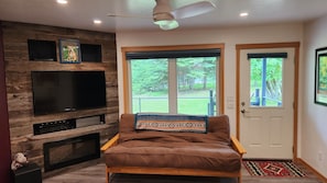 A living room with a large flat-screen TV, a fireplace, a brown leather sofa, and a ceiling fan.