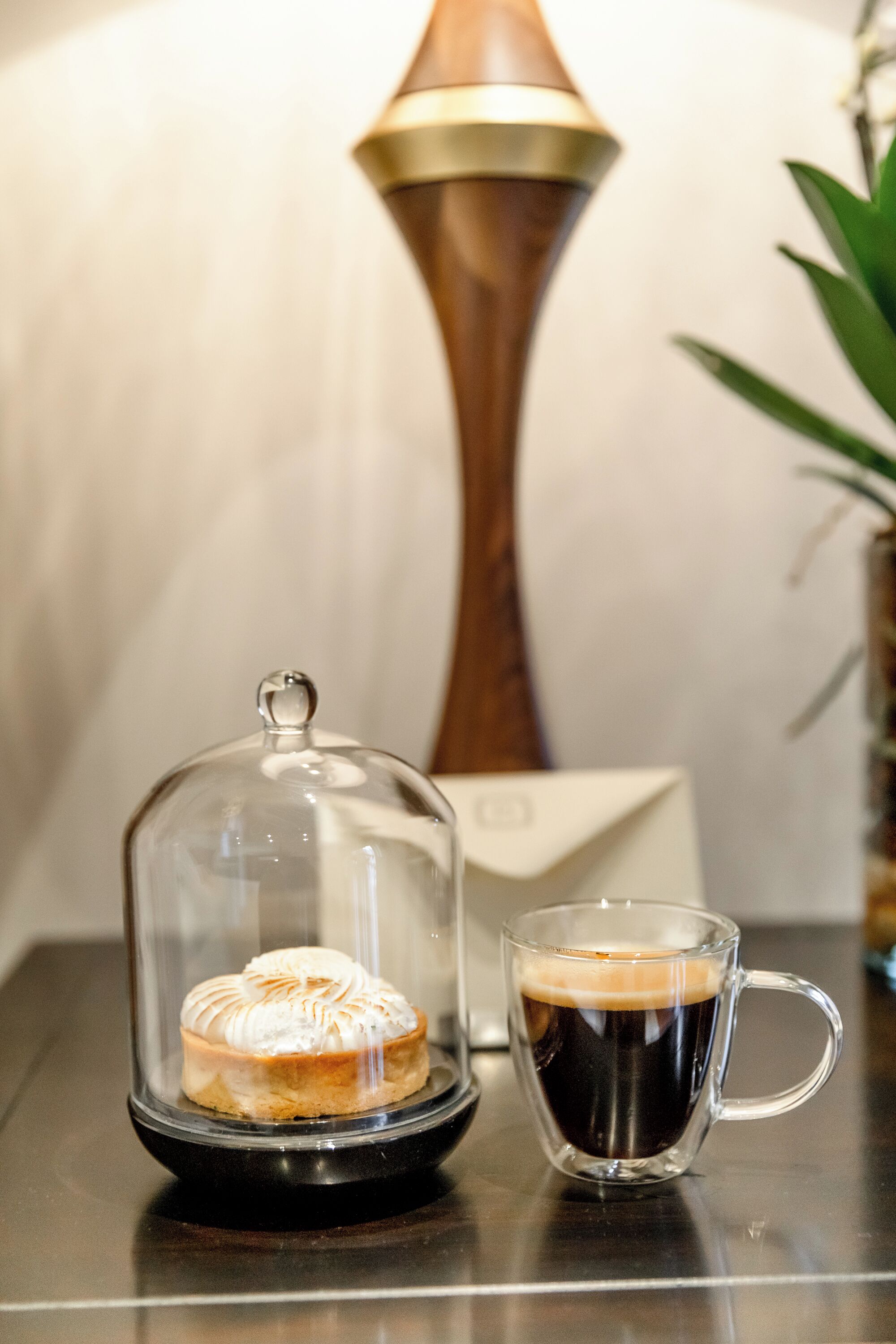 Un dessert présenté sous un cloche de verre, une tasse de café et une lampe sur une table.