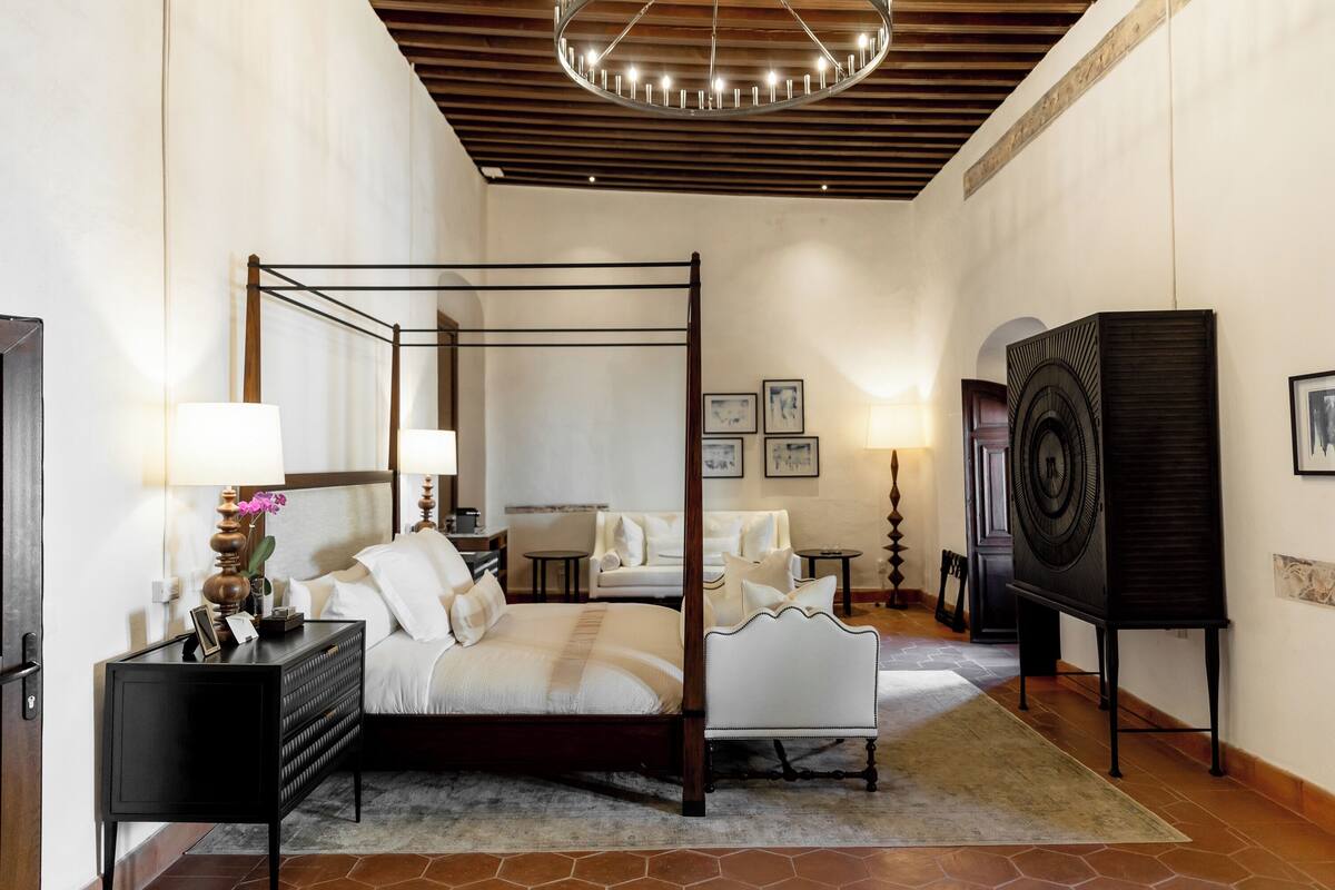 A four-poster bed in a hotel room with a wooden ceiling, a black nightstand, a lamp, and a framed picture on the wall.