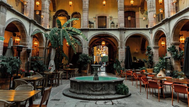 Un patio amplio con varias mesas y sillas para comer, una fuente central y una serie de arcos.