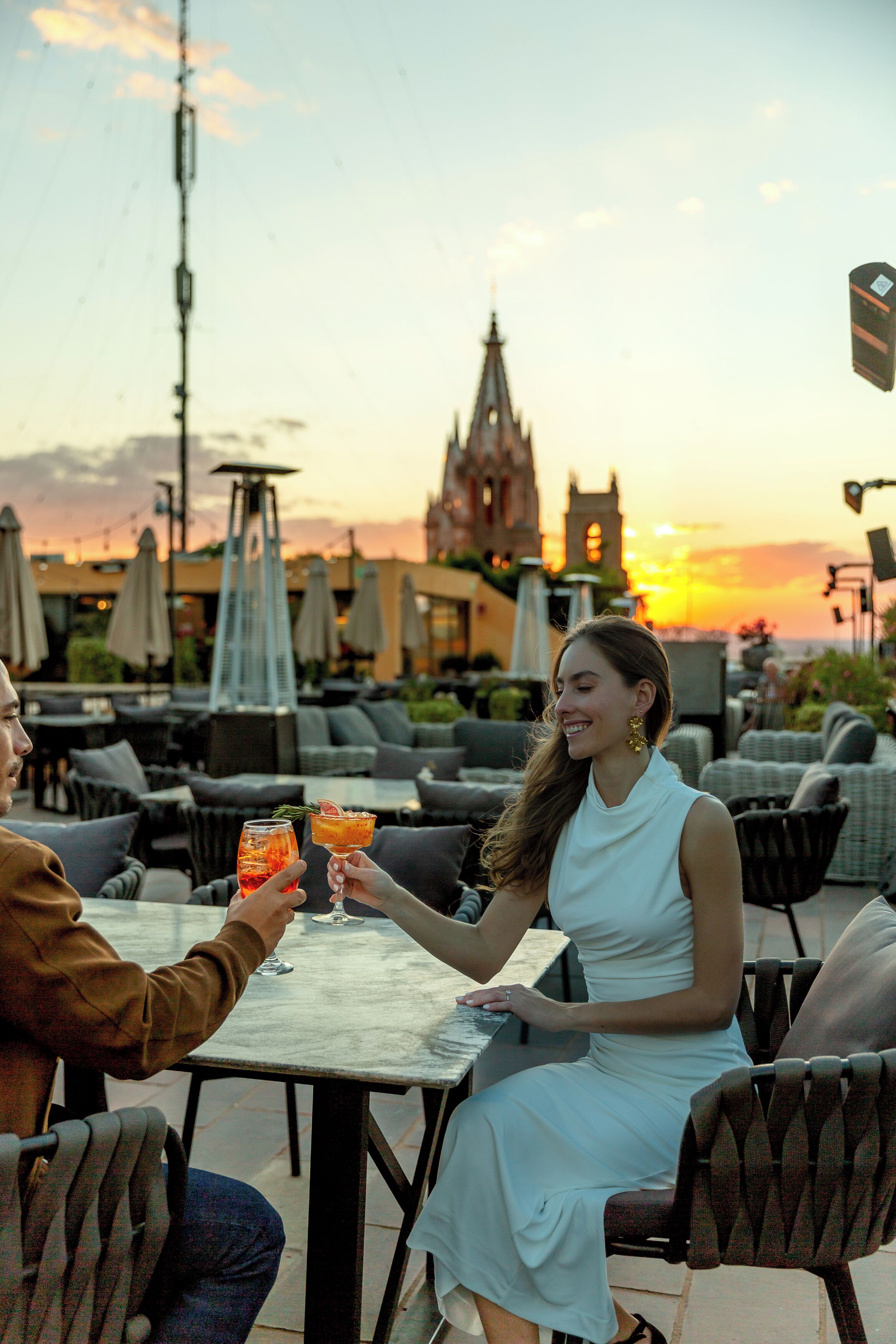Een stel zit aan een buitentafel met drankjes, met uitzicht op een stadsbeeld met een kerktoren en zonsondergang.