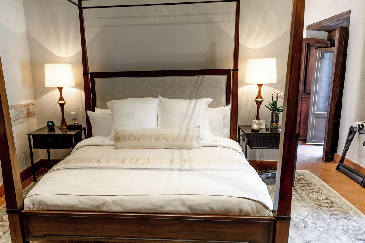 A four-poster canopy bed with white linens, two bedside tables with lamps, and a wooden floor.