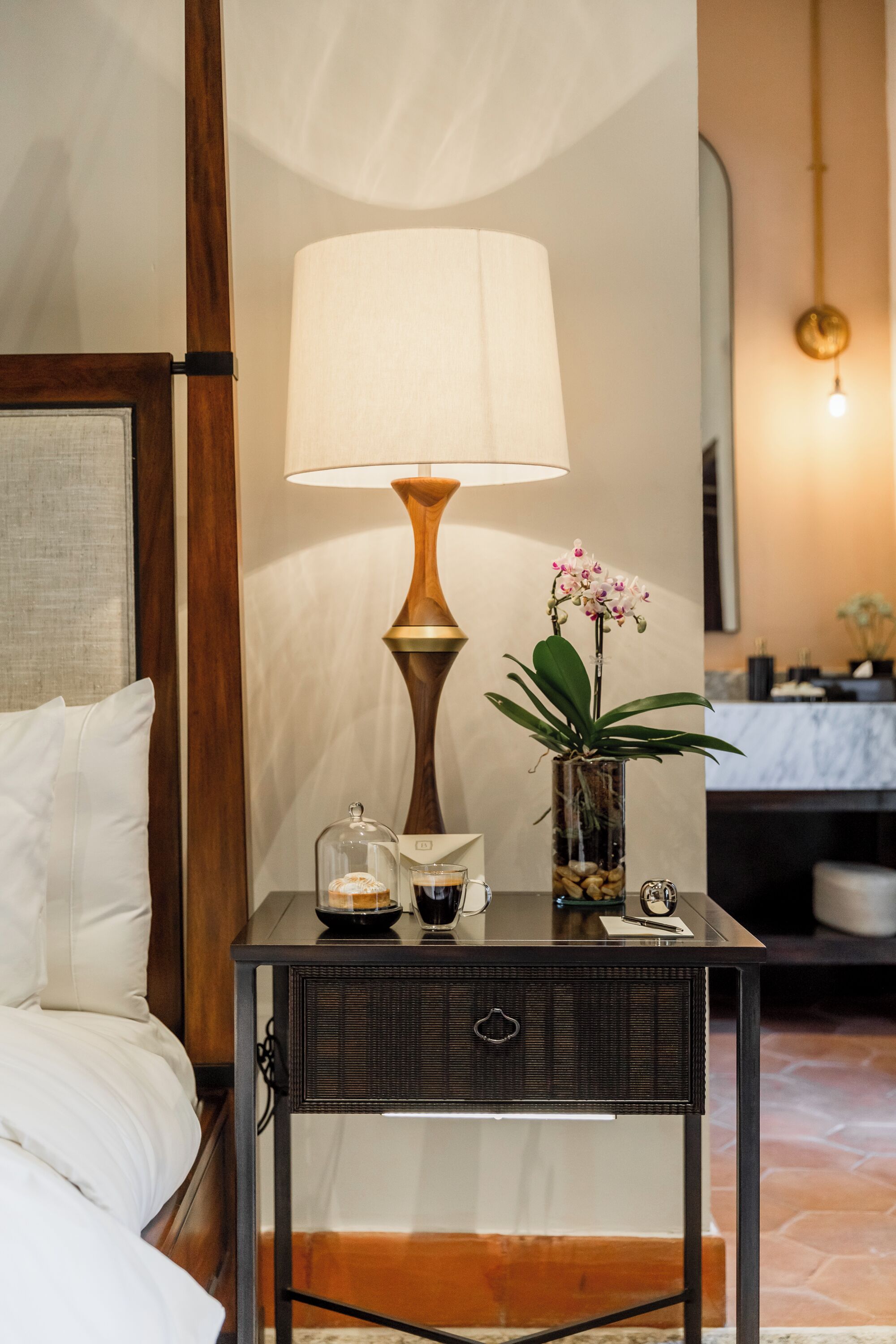 A neatly arranged hotel room with a bed, a bedside table with a lamp, a vase with flowers, and a small decorative item.