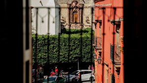 Vista de un edificio histórico con una torre de campanas y una cúpula, rodeado por un patio con vegetación y un edificio rojo.