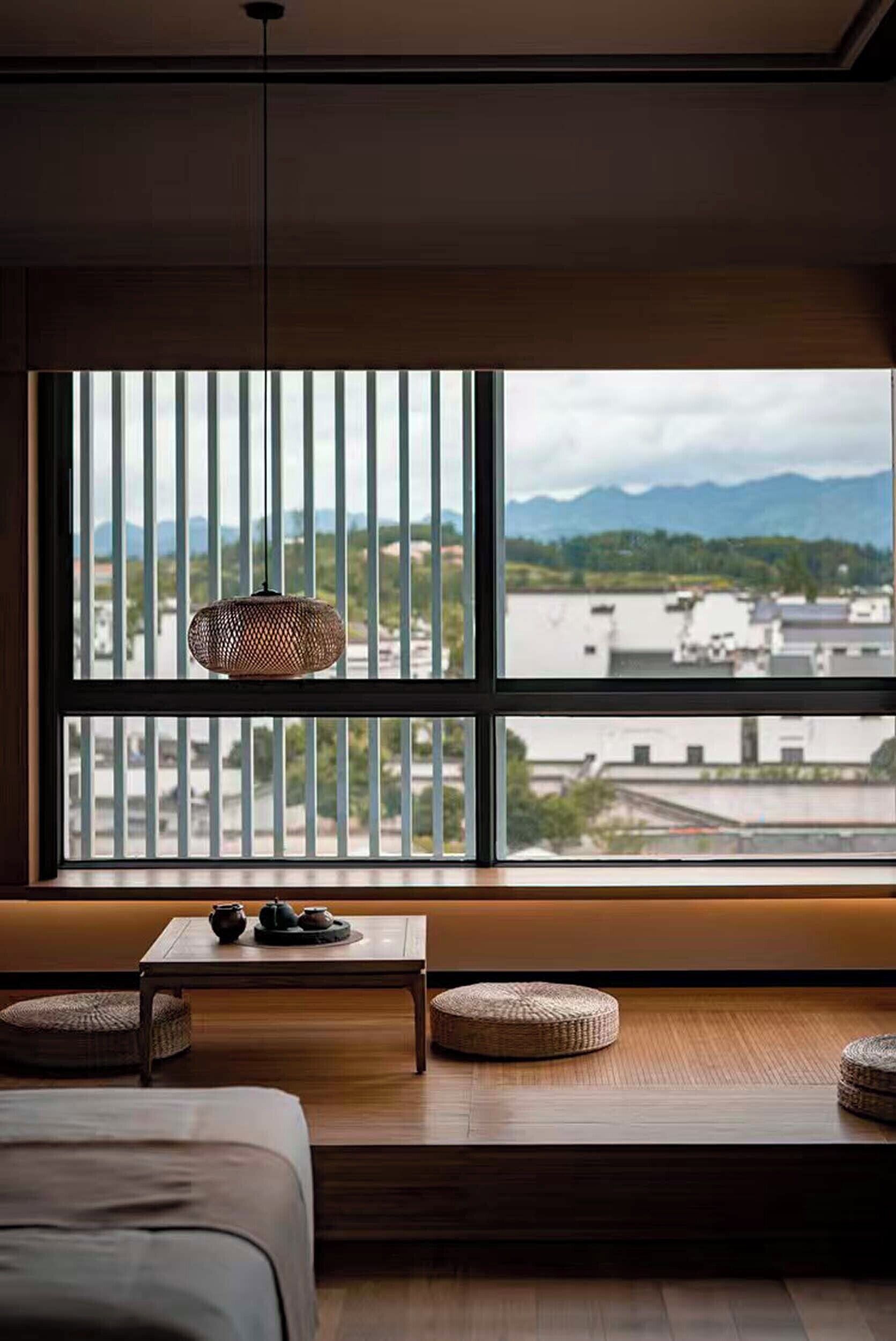 A room with a large window, a hanging light fixture, a small table with a teapot, and a sofa.