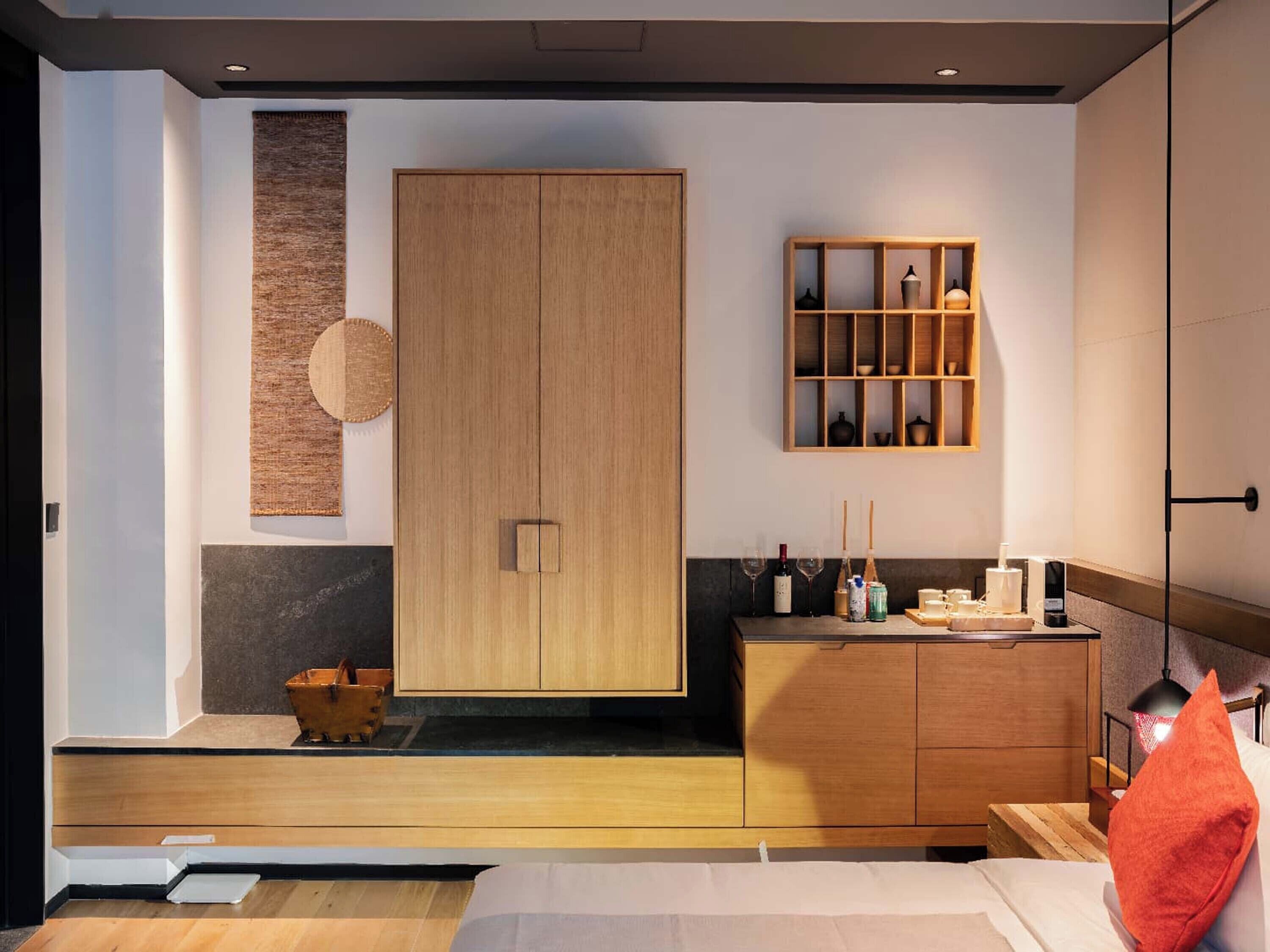 A modern bedroom with a wooden wardrobe, a bedside table with a lamp, and a small shelf with decorative items.