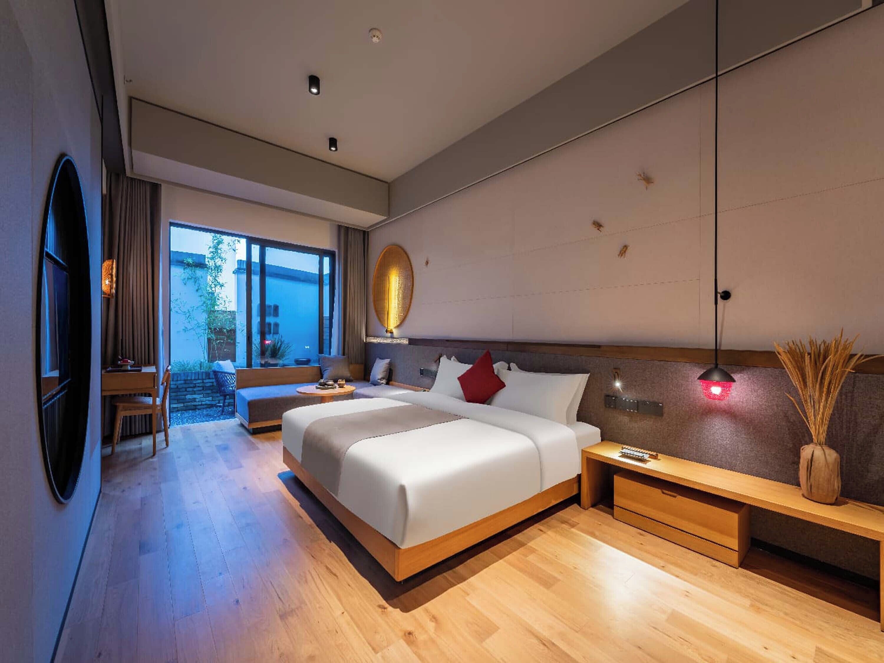 Una habitación de hotel moderna con una cama grande, una mesita de noche, un escritorio y una zona de estar con vistas al exterior.