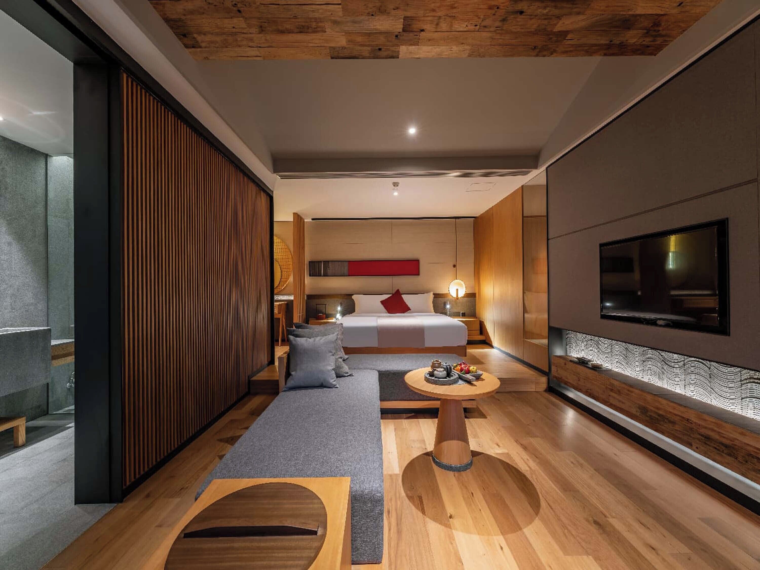 A modern hotel room with a wooden ceiling, a large bed, a TV mounted on the wall, and a small round table with a tray of fruit.
