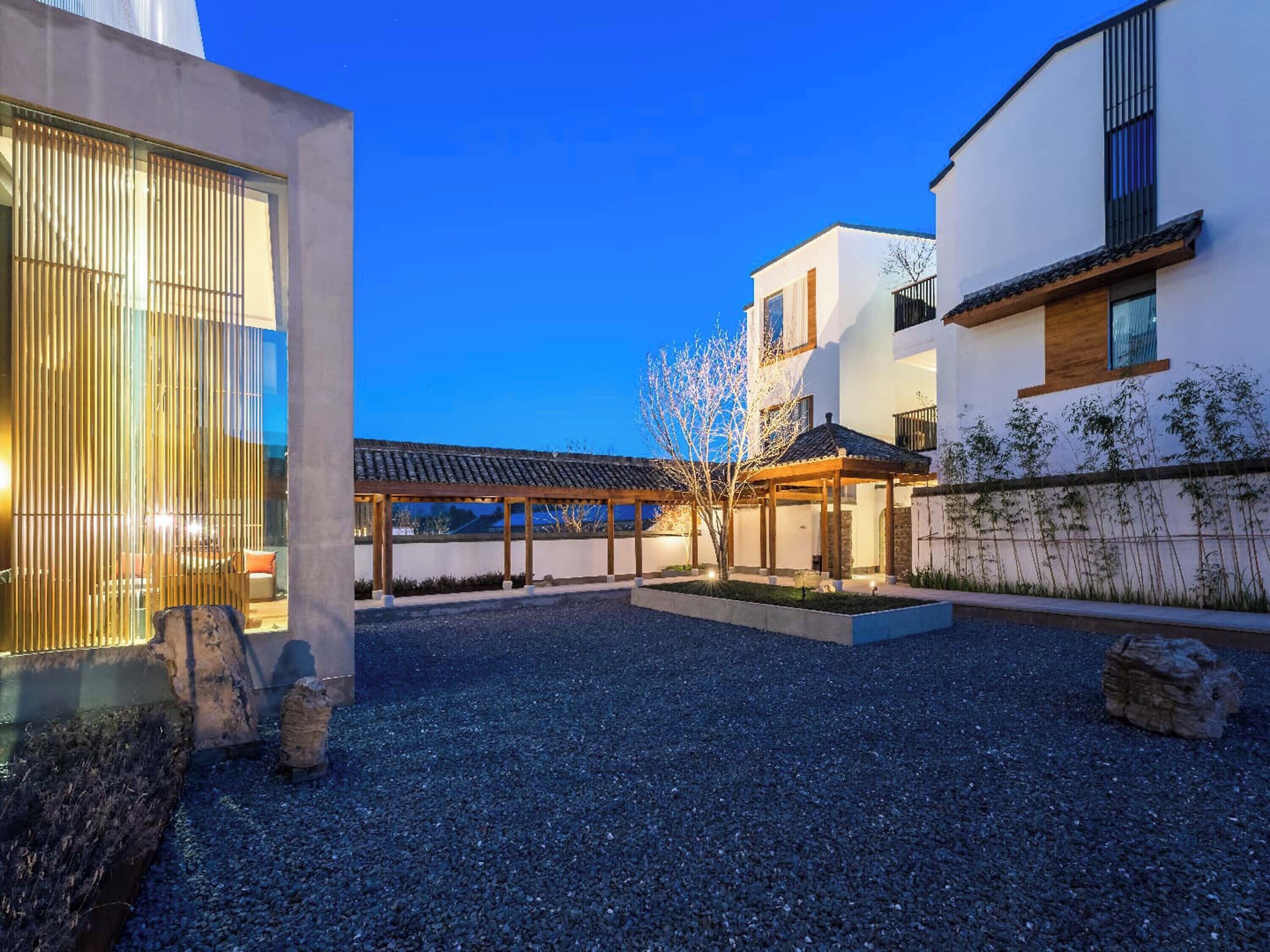 Ein moderner Außenbereich mit einem Kieshof, einer überdachten Sitzgelegenheit und einem Gebäude mit weißer Fassade und Holzdetails.