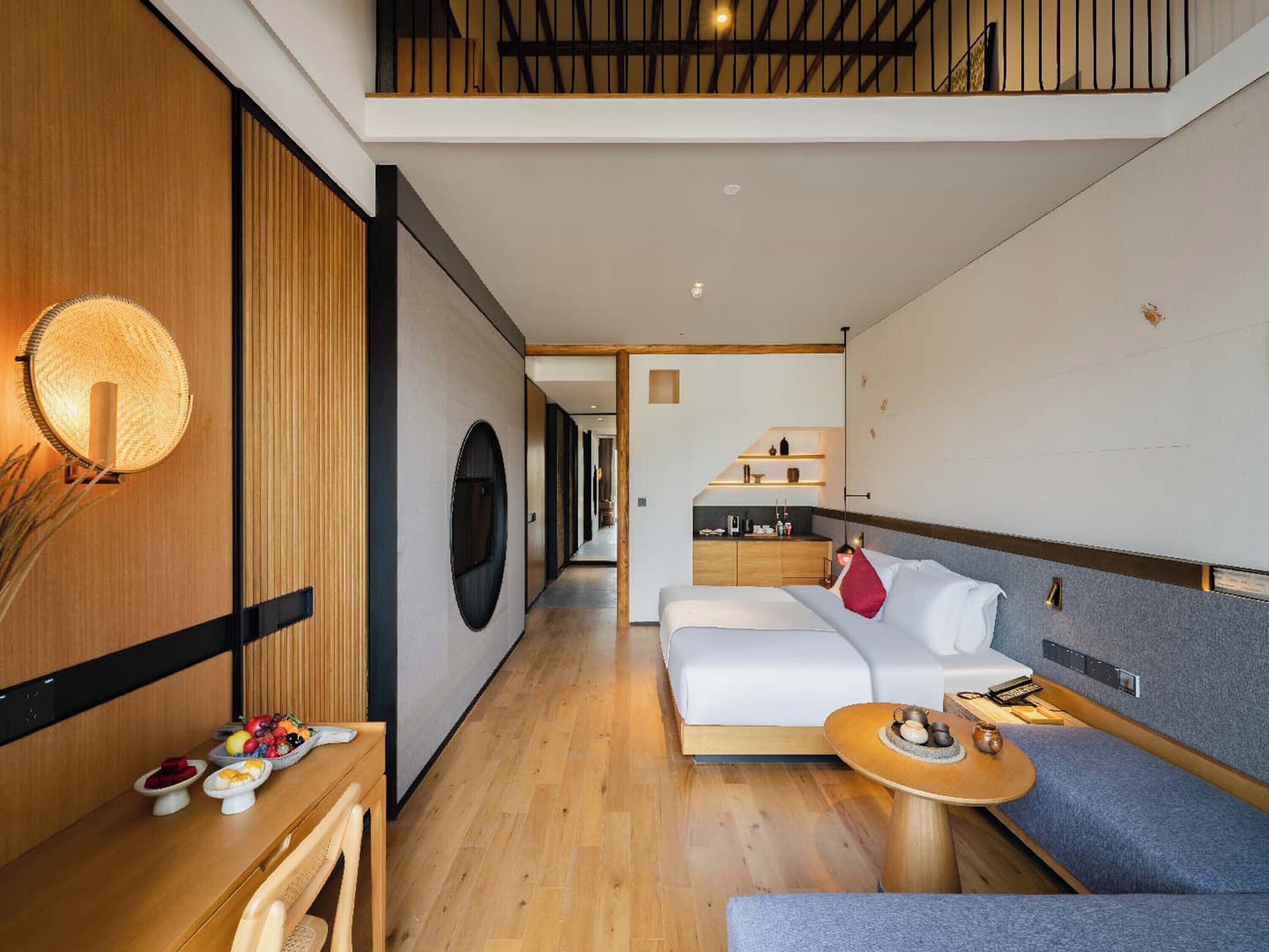 A modern hotel room with a wooden floor, a sofa, a dining table with fruit, and a kitchen area in the background.