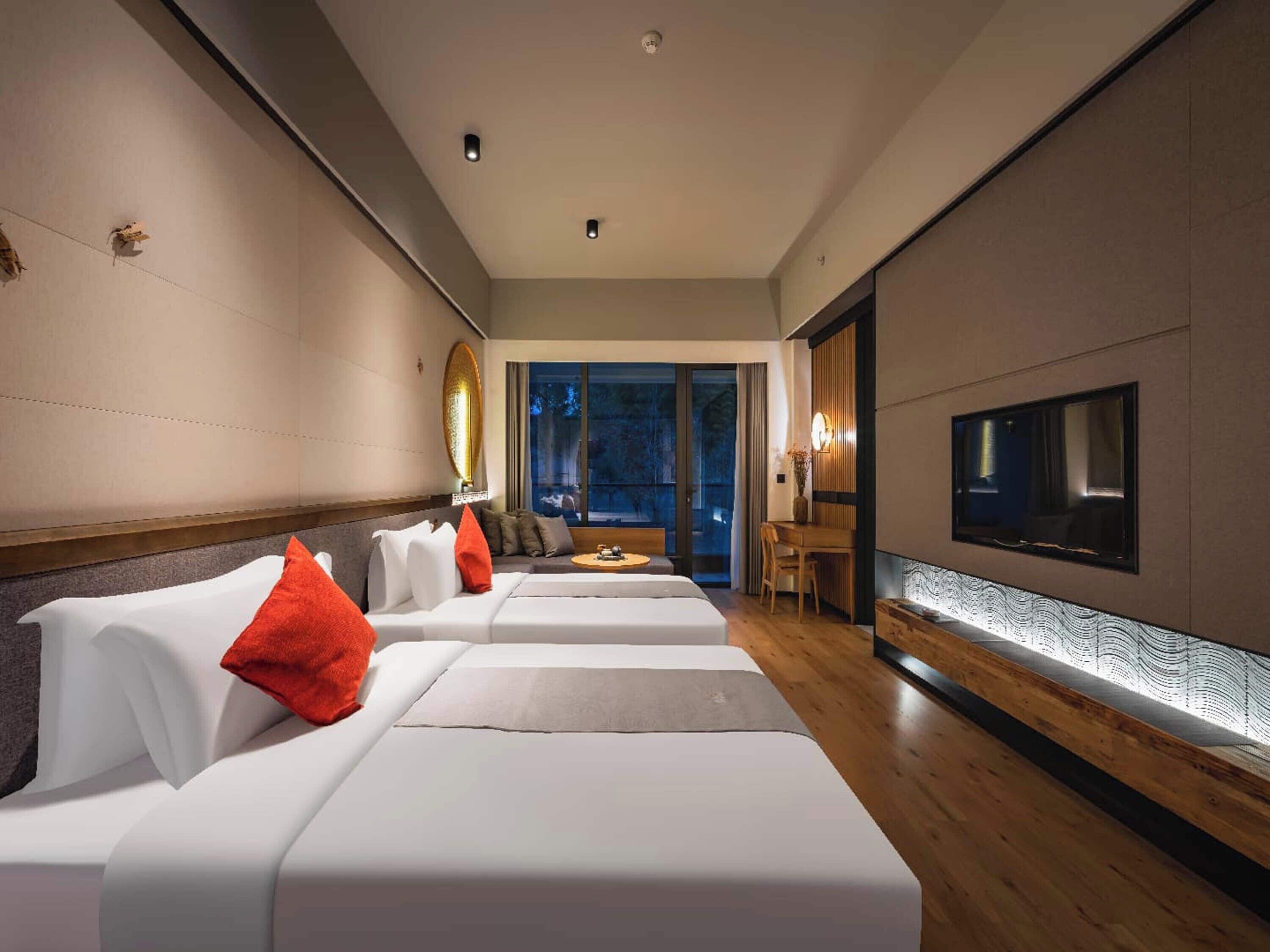 A modern hotel room with two white beds, a wooden floor, a large TV, and a view of the outdoors.