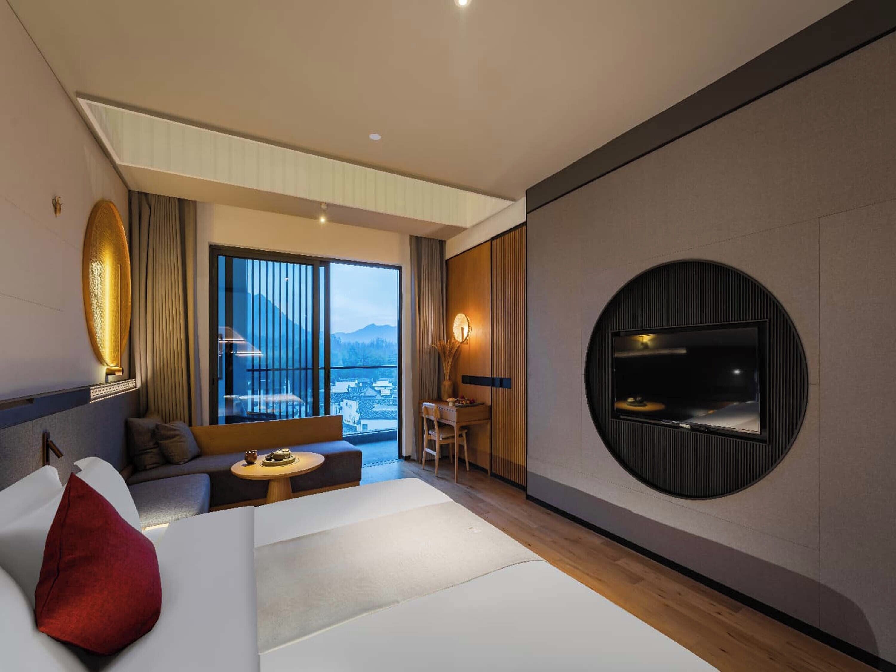 A modern hotel room with a large bed, a TV mounted on the wall, a balcony with a view of the city, and a seating area with a table and chairs.