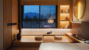 A modern hotel room with a large window offering a view of mountains and trees. There is a bed, a small table with a lamp, and a shelf with decorative items. - Tan She Tang Quan Resort Hotel-Huangshan (Huangshan)