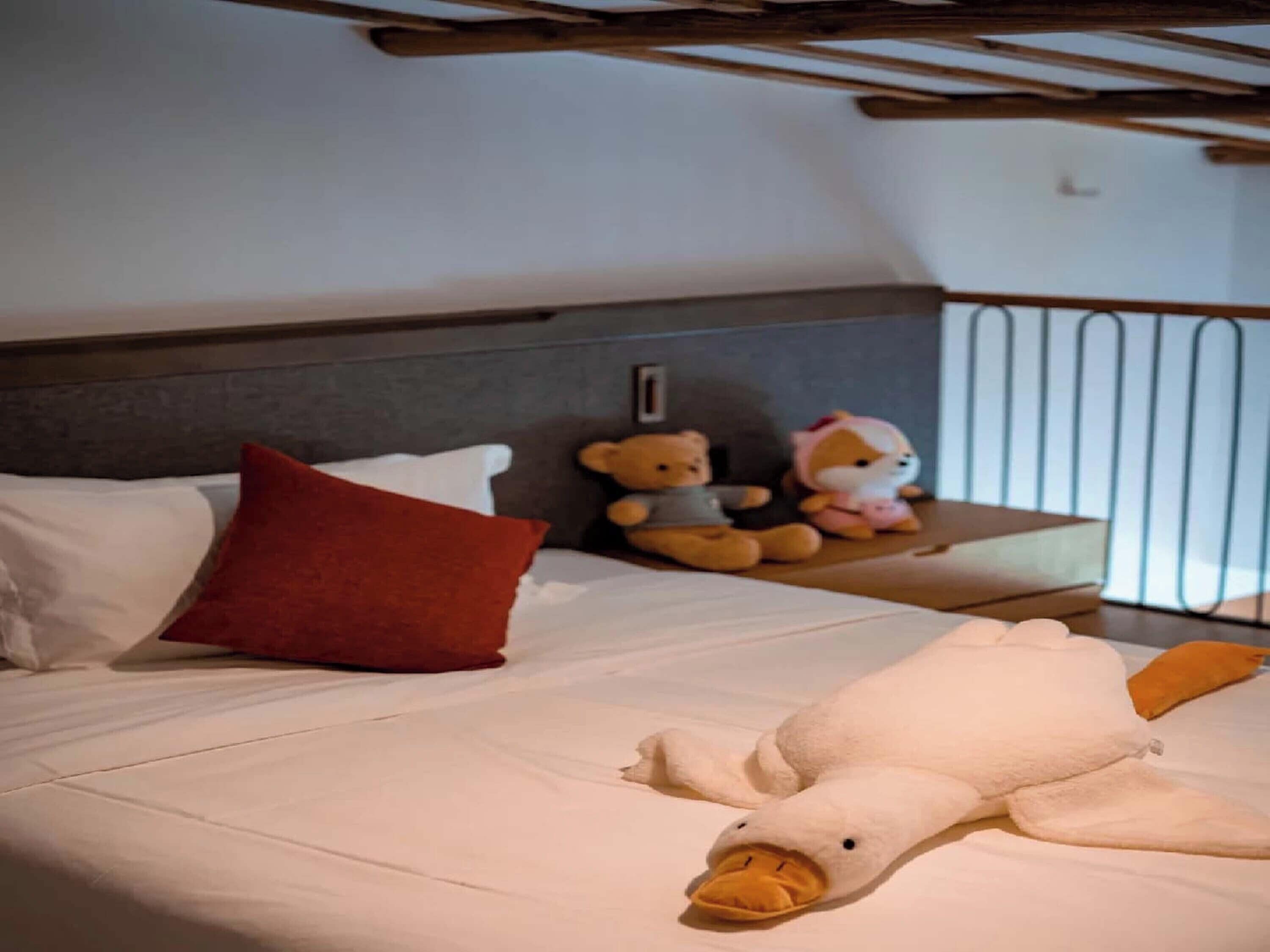 A bed with white bedding, a red pillow, and two stuffed animals on a nightstand.