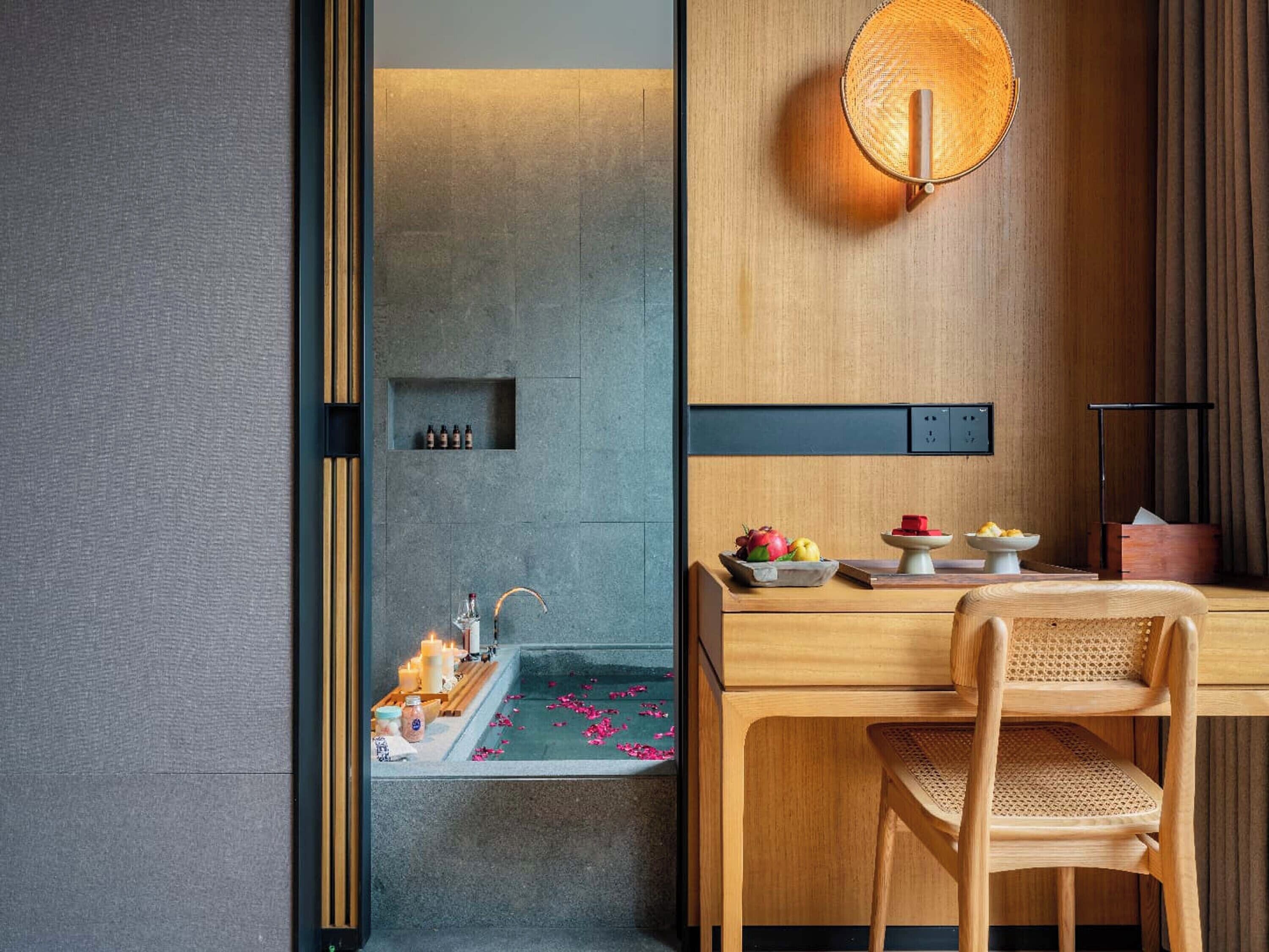 Una habitación de hotel moderna con una mesa de comedor de madera, una lámpara de fibra y un pequeño altar con velas y flores.