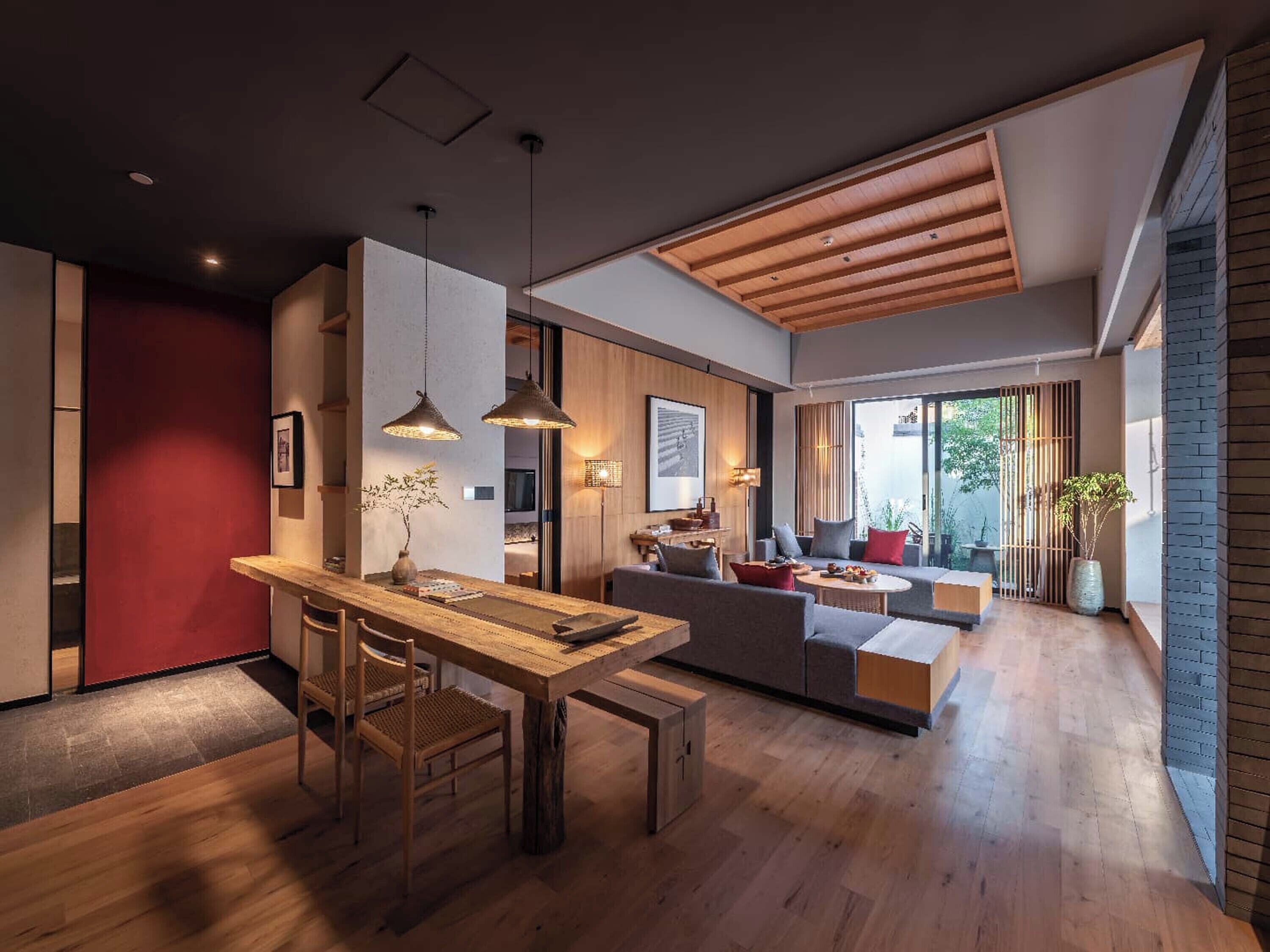 A modern interior with a wooden dining table, chairs, a sofa, and a bench. There are pendant lights, a red accent wall, and a large window with a view.