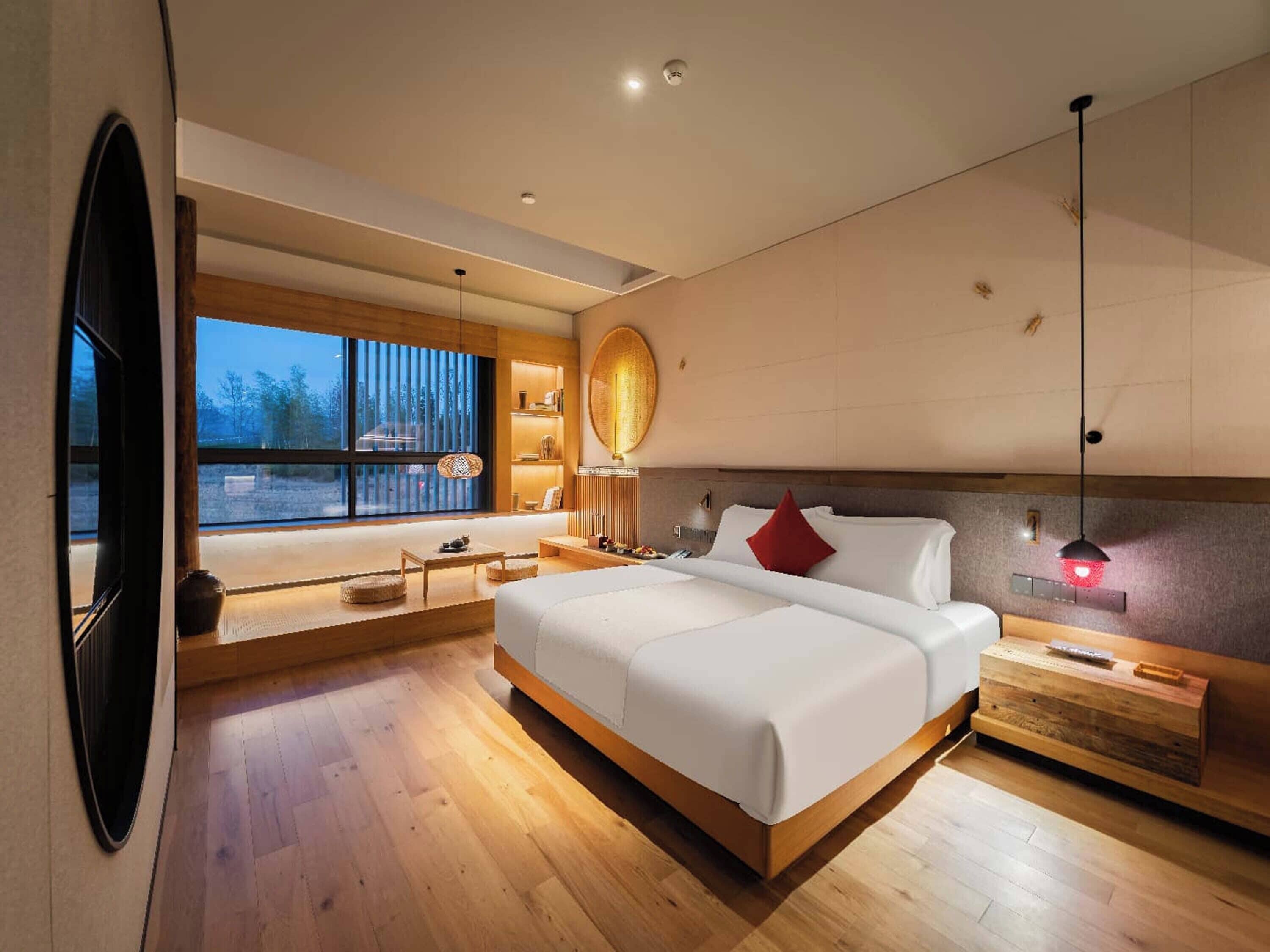 A modern hotel room with a large bed, wooden flooring, a view through large windows, and a cozy seating area with a lamp.