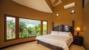 A bedroom with a large bed, a nightstand, and a window with a view of greenery. - Amazing Mountain & Ocean Views (Playa Hermosa)