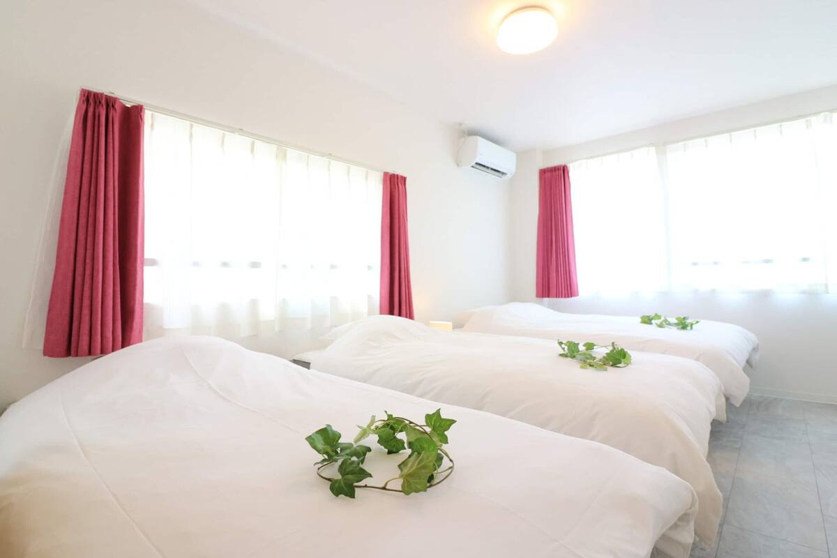 Habitación de hotel con dos camas, cada una con ropa blanca y una planta sobre la cama. Cuenta con ventanas amplias con cortinas rojas y un equipo de aire acondicionado en la pared.