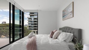 Une chambre moderne avec un grand lit, une tête de lit grise et une vue sur le paysage urbain grâce à de grandes fenêtres du sol au plafond.