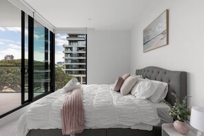 A modern bedroom with a large bed, a grey headboard, and a view of a cityscape through floor-to-ceiling windows. - Free parking - trackside apartment with pool, gym (Moonee Ponds)