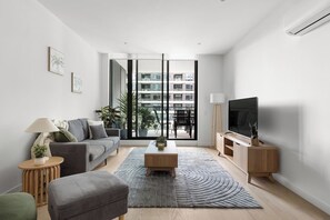 Un salon moderne avec un canapé, une table basse et un meuble pour la télévision. Une grande fenêtre offre une vue sur les bâtiments environnants.