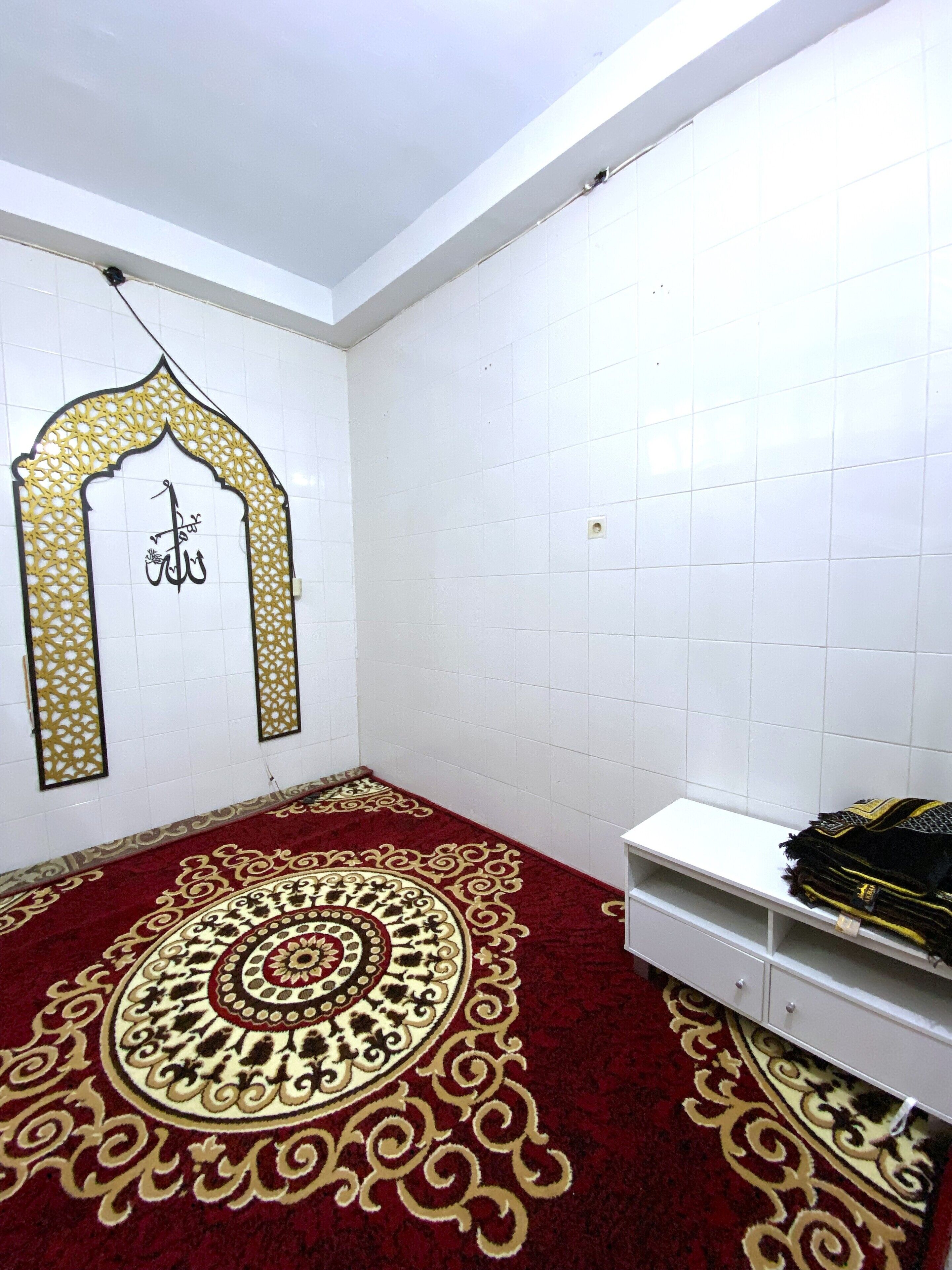 A room with white tiled walls, a decorative carpet with intricate patterns, a white shelf, and a wall-mounted decorative piece with Arabic calligraphy.