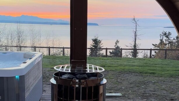 Un bain à remous et un poêle à bois se trouvent sous une terrasse couverte, offrant une vue sur l’océan au coucher du soleil.