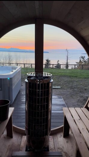 A hot tub and a wood-burning stove under a covered deck with a view of the ocean at sunset. - Whidbey Island Retreat  View, Hot tub, Sauna, EV (Coupeville)