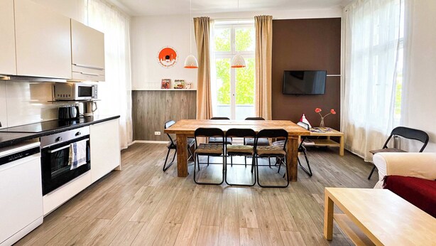 A modern kitchen with white cabinets, a wooden dining table, and a TV mounted on the wall.