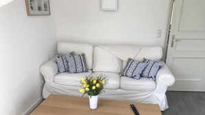 Een woonkamer met een witte bank, gekleurde kussens, een houten salontafel en een vaas met gele bloemen.