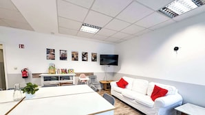Una habitación moderna y luminosa con una mesa blanca, sillas, un sofá blanco con cojines rojos, un televisor montado en la pared y un extintor.