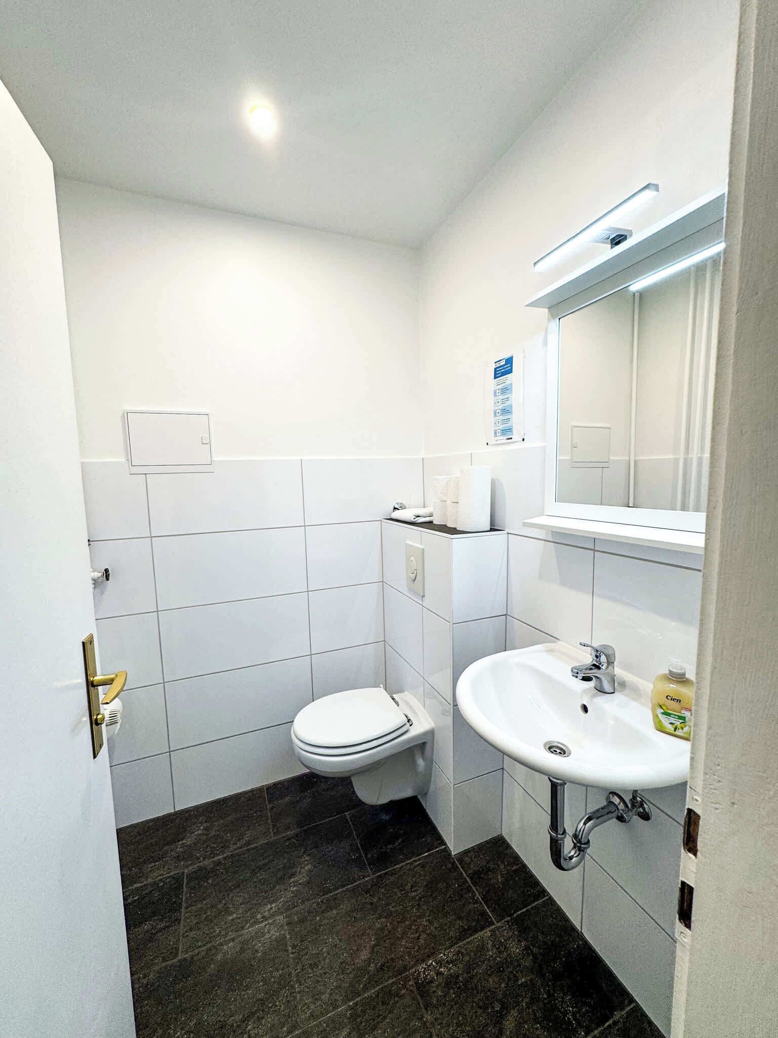 Et lille, rent badeværelse med hvide fliser, et hvidt toilet, en håndvask med spejl og mørkt gulv.