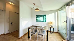 Comedor con una mesa de madera, sillas blancas y una lámpara colgante. Hay una puerta de vidrio que da a un balcón.