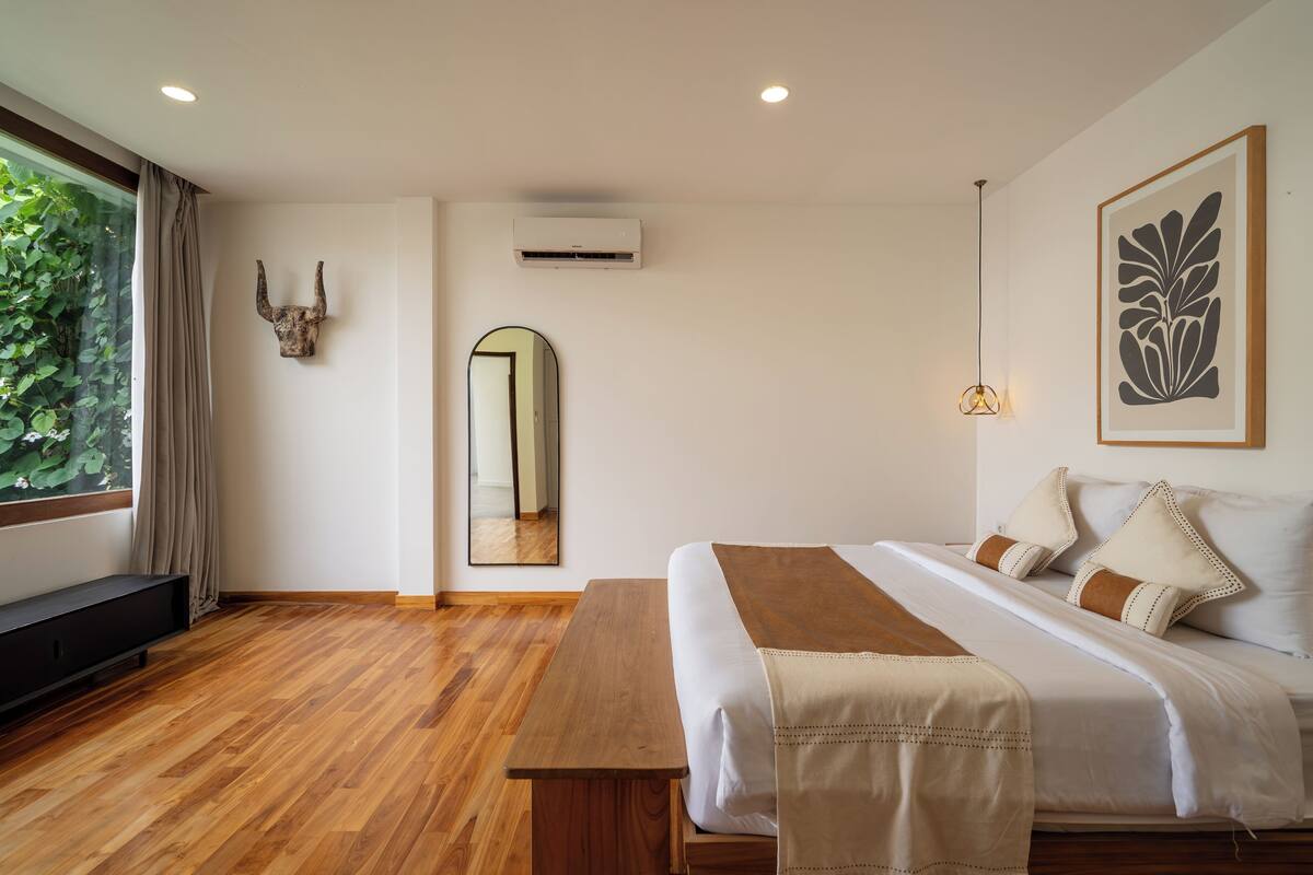Quarto moderno com piso de madeira, uma cama grande com roupa de cama branca, um banco, um espelho e uma obra de arte emoldurada na parede.