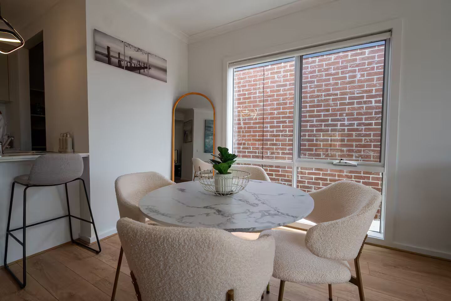 A dining area with a round marble table, four chairs, a plant in a wire basket, and a large window with a brick wall view.