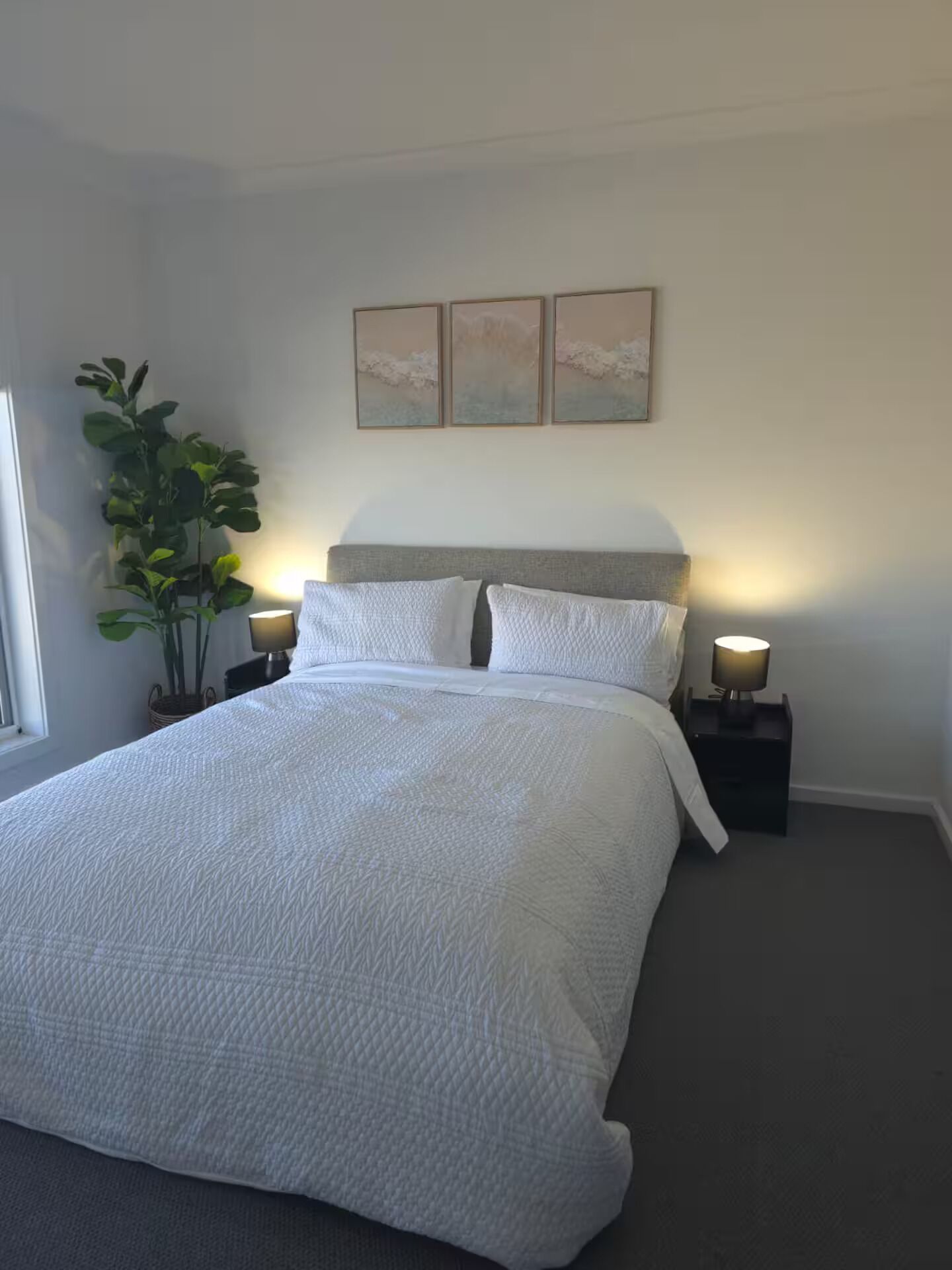 A bedroom with a large bed, two bedside tables with lamps, a potted plant, and three wall-mounted artworks.