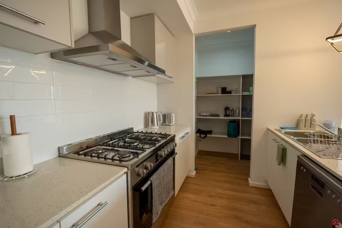 A modern kitchen with a gas stove, stainless steel appliances, and a built-in oven. There is a pantry with shelves and a dish rack.