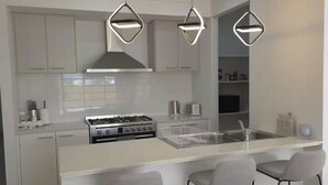 Una cuina moderna amb armaris blancs, una illa central amb tamborets i una placa de gas, amb il·luminació penjant.