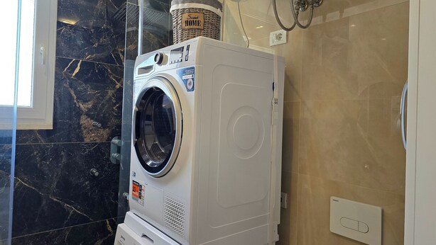 Máquina de lavar e secar empilhadas em um banheiro com parede de mármore e uma cesta com a etiqueta 'Smart Home' por cima.