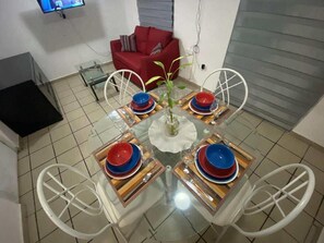Comedor con una mesa de vidrio dispuesta para cuatro personas, rodeada de sillas blancas. Un sofá rojo está al fondo y un televisor montado en la pared.